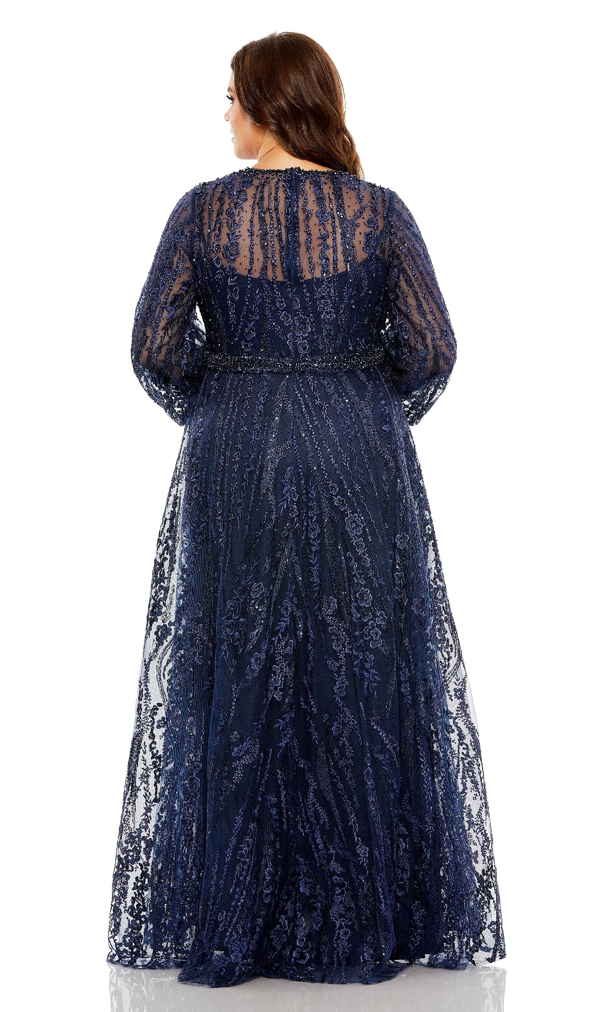 A woman stands facing away, modeling the Mac Duggal 20472— a long plus-size formal dress in navy blue, featuring sheer sleeves, intricate sequin details, and delicate floral embroidery along the v-neckline, set against a plain white background.